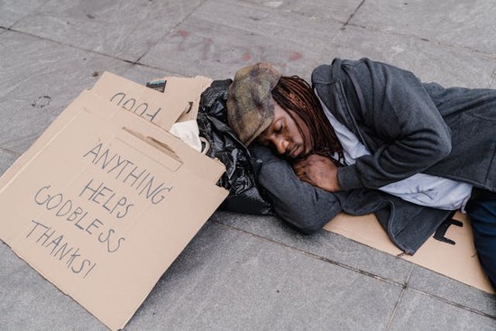 A homeless man lies on the ground beside a cardboard sign that asks for donations.