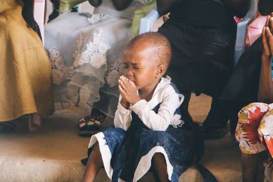 A small child sits on the edge of a step in a church service. She has her small hands folded and her eyes tightly closed. She is sincerely praying to God.