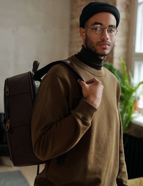 A young man wearing glasses, a hat, and a backpack looks at the camera.