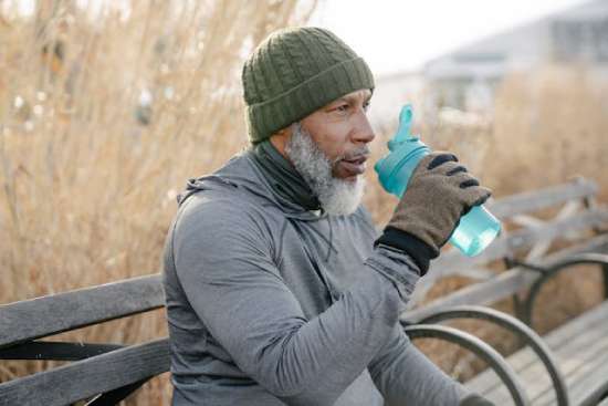 An older man in workout clothing sits outside on a park bench and sips from a shaker bottle. Exercise, hydration, and time outside are all a part of the Adventist health message and a key to longevity.