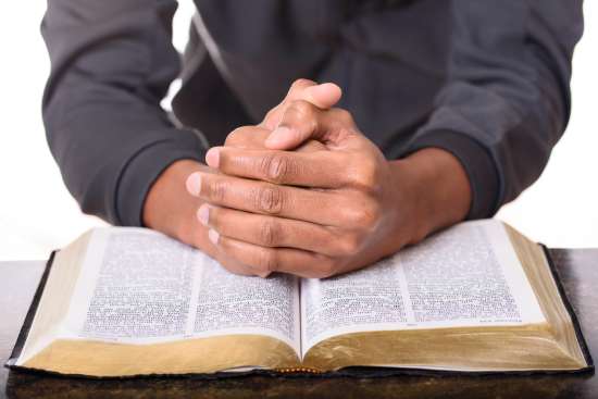 A close up of hands folded in prayer over an open Bible.