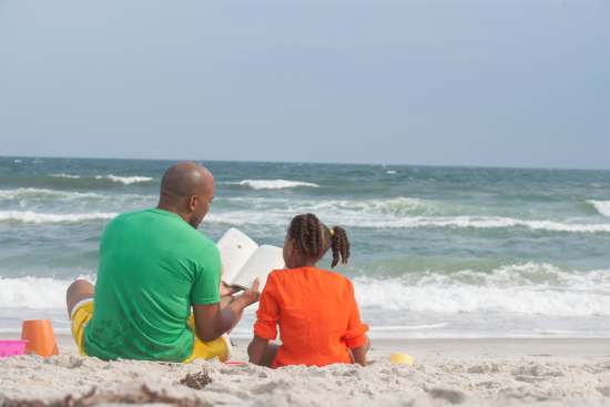 A father sits on the beach with his daughter and reads to her