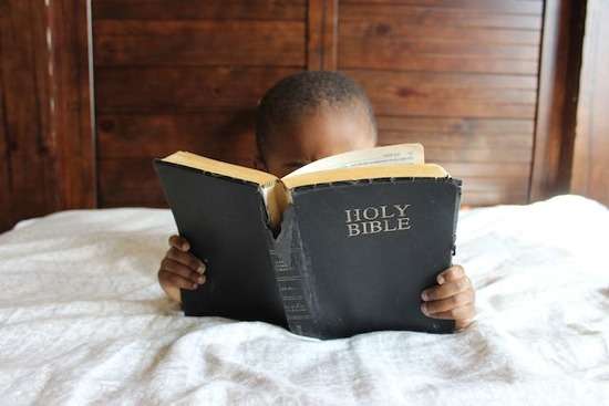A young child lays in a bed with his face buried in the Bible.