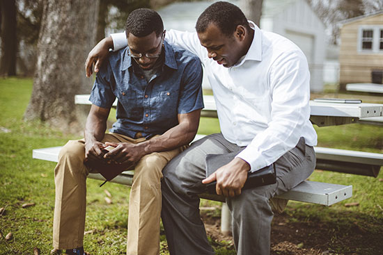 Two men holding Bibles pray together on a park bench
