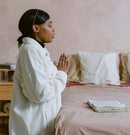 A woman kneels by her bed with folded hands over an open Bible.