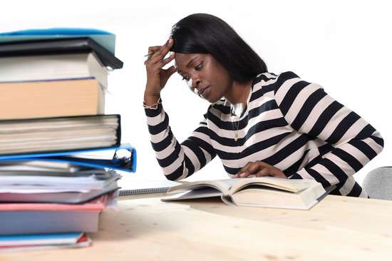 A young woman with her hand to her forehead in exasperation and a large pile of books in front of her seems to struggle to understand what she is reading.